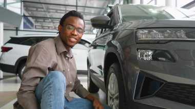 Gülümseyen Afrikalı Amerikalı bir adam galeride yepyeni bir arabanın yanında oturuyor, satın almadan önce dış görünüşünü ve tekerleklerini dikkatlice inceliyor..