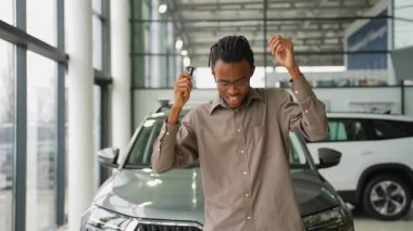 Mutlu Afrikalı Amerikalı adam yeni araba alımını kutlamak için galerinin anahtarlarını tutarken neşeyle dans ediyor..