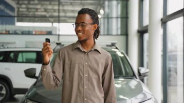 Gülümseyen Afrikalı Amerikalı bir adam araba anahtarlarını bir galeride tutuyor. Yeni aldığı arabayı neşe ve heyecanla kutluyor..