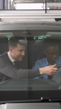 Bir araba satıcısı müşterinin yanında oturur ve satın almadan önce aracın özelliklerini açıklar. Alıcı direksiyonu modern bir araba galerisinde tutuyor..