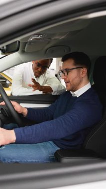 Bir araba satıcısı, sürücü koltuğunda oturan potansiyel bir alıcıya sağdan sürüş aracının iç hatlarını sunar.