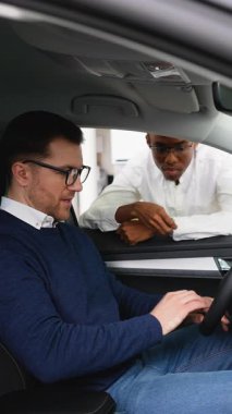 Bir araba satıcısı, bir satış şirketinde sürücü koltuğunda oturan bir müşteriye satın alma kararı vermesine yardımcı olan bir aracın özelliklerini açıklar..