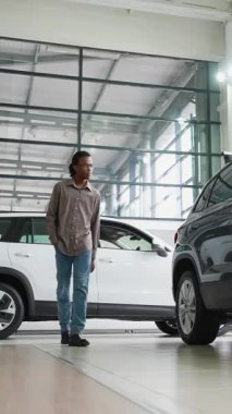 Genç bir adam parlak bir araba galerisine gider, çeşitli araçları inceler ve bir sonraki araba alımı için seçenekleri değerlendirir..