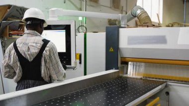 Koruyucu teçhizat kullanan Afrikalı Amerikalı işçi dijital kontroller kullanarak bir mobilya imalat fabrikasında OSB ahşap levhaları kesmek için CNC testere kullanıyor..