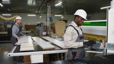 İki fabrika işçisi, lamine ahşap tahtaları mobilya parçalarına ayırmak için bir CNC testeresi kullanıyor. Biri makineyi kontrol ediyor, diğeri de kesik parçaları yığıyor..