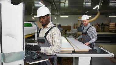 İki fabrika işçisi, lamine ahşap panelleri kesmek için CNC makinesi kullanıyor. Biri dijital kontrolleri yönetirken, diğeri arkaplandaki parçaları keser..