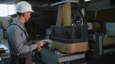 Sert şapkalı fabrika işçisi, bir üretim tesisinde mobilya üretimi için büyük ahşap panelleri küçük parçalara ayırmak için CNC makinesi kullanıyor..
