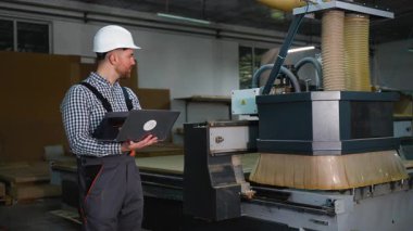 Modern bir mobilya üretim tesisinde otomatik CNC yönlendirici kullanarak tahta panelleri kesen koruyucu ekipman mühendisi..