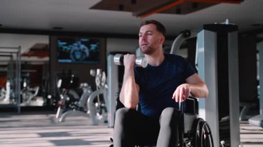 Tekerlekli sandalyede oturan bir adam spor salonunda dambılla biseptik kıvrımlar yapıyor, dayanıklılık, kapsama ve aktif yaşam tarzını teşvik ediyor..