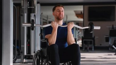 Tekerlekli sandalyedeki adam spor salonundaki kasları çalıştırmak için kıvrımlı çubuğu kaldırıyor, fitness ve adaptif güç oluşturmaya olan bağlılığını gösteriyor..
