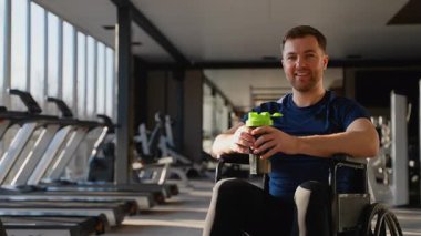 Modern güneşli bir spor salonunda egzersiz yaptıktan sonra elinde protein karıştırıcı tutan tekerlekli sandalyedeki mutlu atletik adam. Fitness ve sağlıklı yaşam tarzı konsepti.