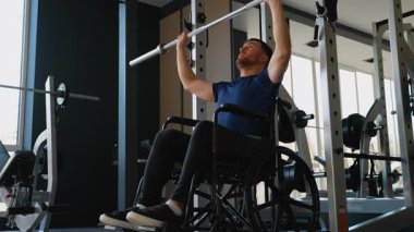 Tekerlekli sandalyede oturan bir adam spor salonunda halter omuz presi yapar. Güç, kararlılık ve fitness hedeflerine bağlılık gösterir..