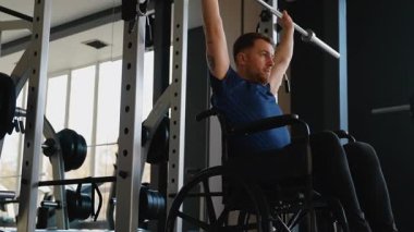 Tekerlekli sandalyedeki odaklanmış adam spor salonunda halter presi yapıyor. Etkinlikte güç eğitimi, motivasyon ve adaptif uygunluk.