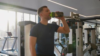 Parlak bir spor salonunda egzersiz yaptıktan sonra protein içen atletik bir adam, egzersiz sonrası iyileşme ve beslenmeyi destekliyor..
