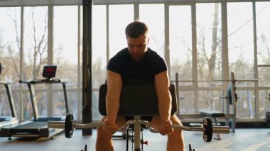 Atletik bir adam iyi ışıklandırılmış bir spor salonunda EZ barla biceps vaazı veriyor. Düzgün bir forma ve kas kasılmasına konsantre oluyor..