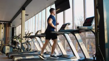 Geniş pencereli, parlak ve modern bir spor salonunun içinde koşu bandında koşan genç adam, kardiyo fitness ve dayanıklılık eğitimine odaklanıyor..