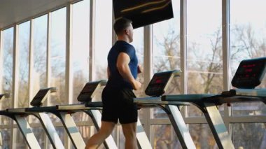 Modern bir spor merkezinde koşu bandında koşan atletik bir adam, sabah güneşi büyük pencerelerden içeri giriyor. Kalp ve sağlıklı yaşam tarzı kavramı.
