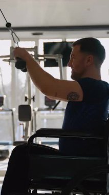 Tekerlekli sandalyedeki gülümseyen adam spor salonundaki kablo makinesinde vücudunun üst kısmını eğitir. Kararlılık, güç ve adaptasyon rutini gösterir..