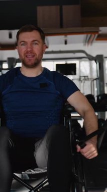 Tekerlekli sandalyedeki adam spor salonunda çift kablo makinesiyle çalışıyor, üst vücut gücünü arttırıyor ve adaptasyon ve motivasyon gösterisi yapıyor..