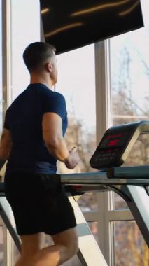 Modern bir spor merkezinde koşu bandında koşan atletik bir adam, sabah güneşi büyük pencerelerden içeri giriyor. Kalp ve sağlıklı yaşam tarzı kavramı.