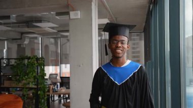 Gülümseyen Afrikalı Amerikalı erkek akademik önlük ve şapka ile mezun olduktan sonra modern üniversite koridorlarında yürür ve eğitimini tamamlar..