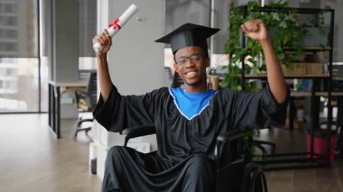 Tekerlekli sandalyedeki neşeli Afrikalı Amerikalı öğrenci mezuniyeti kutluyor, gülümsüyor ve diplomayla kollarını kaldırıyor..