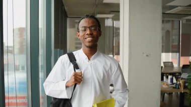 Kendine güvenen Afrikalı Amerikalı tıp öğrencisi ya da stajyer elinde kitaplarla modern bir üniversite koridorunda yürürken gülümsüyor..