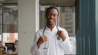 Boynunda steteskopla modern bir klinik koridorunda ya da lobide güvenle duran beyaz önlüklü mutlu Afrikalı genç bir doktor..