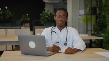 Masasında dizüstü bilgisayar ve steteskopla oturan, modern ofiste tıbbi notlar yazan, beyaz önlüklü, odaklanmış Afrikalı Amerikalı bir doktor..