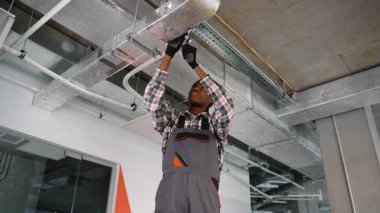 Modern bir ofis binasında tavana monte edilmiş havalandırma kanalı üzerinde çalışan işçi tulumu ve eldivenli Afrikalı Amerikalı teknisyen..