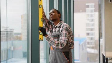 Modern bir binadaki büyük bir pencerenin dikey hizasını kontrol etmek için ruh seviyesini kullanan Afrikalı Amerikalı inşaat işçisi.