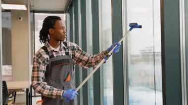 Profesyonel Afrikalı Amerikalı temizlikçi eldiven ve tulum giyer. Parlak, modern ofis alanının camlarını silmek için cam sileceği kullanır..