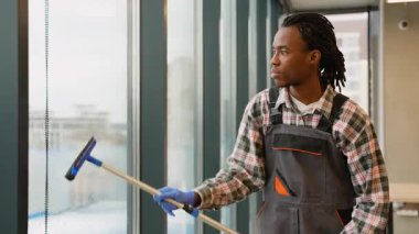 İş üniformalı ve eldivenli bir hademe modern ticari ofis ortamında uzun bir pencereyi temizlemek için bir cam sileceği kullanır..