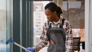 Genç, profesyonel üniformalı hademe, çalışma saatlerinde modern bir ofis binasının camını silmek için cam sileceği kullanıyor..