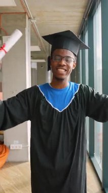 Mezuniyet elbisesi içindeki neşeli Afrikalı Amerikalı öğrenci, akademik başarısından sonra gururla gülümseyen ve kutlama için elini kaldırdı..