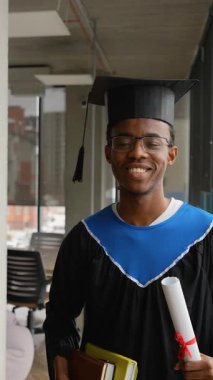 Gülümseyen Afrikalı Amerikalı erkek akademik önlük ve şapka ile mezun olduktan sonra modern üniversite koridorlarında yürür ve eğitimini tamamlar..