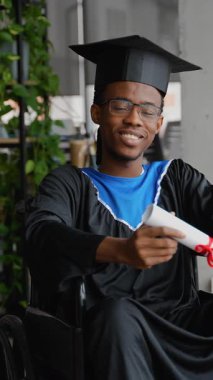 Gururlu Afrikalı Amerikalı mezun, tekerlekli sandalyede oturuyor, akademik önlük ve şapka giyiyor, üniversite mezuniyetinden sonra diplomasını içeride tutuyor..