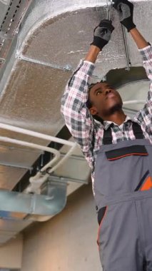 Üniformalı yetenekli Afrikalı Amerikalı teknisyen havalandırma kanalını havalandırma ve havalandırma kanallarını havalandırma ya da bakım işleri sırasında aletlerle sıkıyor..