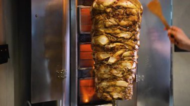 Ticari bir mutfakta ahşap bir spatulayı elinde tutan şefin eliyle dikey bir rostoda pişen büyük bir et yığını..