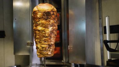Ticari bir mutfak ortamında yanan ocaklarla birlikte dikey bir tükürükte kızaran büyük bir shawarma et yığınının tam görüntüsü.