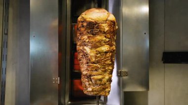 Ticari bir mutfak ortamında yanan ocaklarla birlikte dikey bir tükürükte kızaran büyük bir shawarma et yığınının tam görüntüsü.