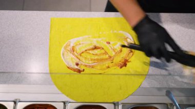 Fast food tezgahında siyah eldiven ve maşa kullanarak shawarma dürümü hazırlamak için sarı ekmek üzerine sos serpiştiren bir şefin tepeden aşağı bakışı..