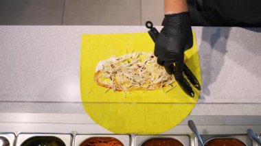 Bir şefin, yemek tezgahında maşa ve eldiven kullanarak soslu ve doğranmış sebzeli sarı ekmek üzerinde donör kebabı bir araya getirirken tepeden aşağı bakışı..