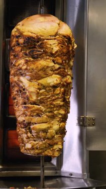 Ticari bir mutfak ortamında yanan ocaklarla birlikte dikey bir tükürükte kızaran büyük bir shawarma et yığınının tam görüntüsü.