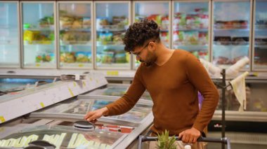 Kahverengi kazaklı Arap bir süpermarkette kırmızı balık ya da somon toplamak için dondurucu açar. Market arabasını iterken dikkatlice deniz ürünlerini seçer..