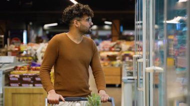 Kahverengi kazaklı bir Arap süpermarketin soğutmalı bölümünün yanında duruyor. Bıldırcın yumurtası almak için rafları dikkatle tarıyor..