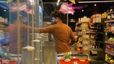 Kahverengi kazaklı bir Arap süpermarkette cam buzdolabının kapısını açar. Market arabasıyla alışveriş yaparken köpüklü içkilere uzanır..