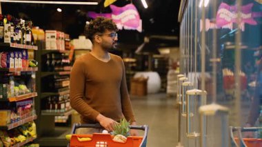 Kahverengi kazaklı Arap bir markette cam bir buzdolabının önünde duruyor, bir alışveriş arabasını iterken özenle köpüklü içecekleri seçiyor..