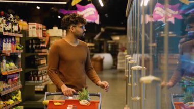 Kahverengi kazaklı bir Arap markette soğutuculu bir bölümde duruyor, soğuk içeceklere göz gezdirirken bir alışveriş arabasını itiyor..