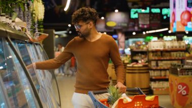 Kahverengi kazaklı bir Arap süpermarkette buzdolabının kapısını açar. Peynir seçerken, içinde taze yiyecek ve süt ürünleri olan bir arabayı iterken..
